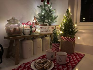 A kitchen counter with jars of tea coffee and sugar a Santa mug with candy canes two small Christmas trees a plate of cookies and two mugs on a red snowflake cloth at Haul A Gwynt in Criccieth