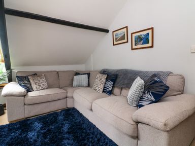 A sitting room with a beige corner sofa with cushions a dark blue rug and framed pictures on the wall at Horseshoe Cottage in Hartland