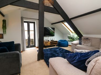 A living room with gray sofas blue cushions a blue bean bag a wooden TV stand black beams and a skylight window at Horseshoe Cottage in Hartland