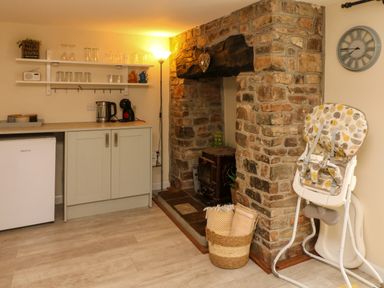 A kitchen area with shelves holding glasses a kettle a coffee machine a fridge a stone fireplace with wood burner a basket with blankets and a high chair at Horseshoe Cottage in Hartland
