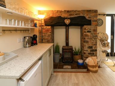 A kitchen area with open shelves holding glasses a kettle and coffee machine countertop a wood burning stove set in a brick fireplace and a baby high chair at Horseshoe Cottage Hartland