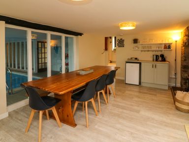 A dining area with a wooden table and black chairs next to a kitchenette and an indoor pool visible through glass doors at Horseshoe Cottage in Hartland