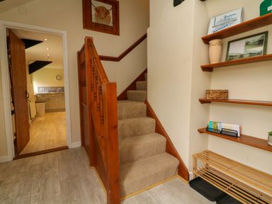 A hallway with a carpeted staircase and wooden shelving next to a doorway leading to a kitchen at Horseshoe Cottage in Hartland