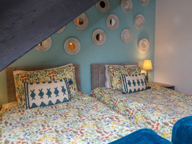 A bedroom with two single beds with floral bedding and decorative pillows under a slanted ceiling with round mirrors on a blue wall at Horseshoe Cottage in Hartland