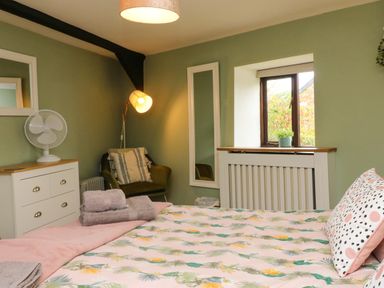 A bedroom with a bed with floral bedding a green armchair a chest of drawers with a fan and a window with a plant at Horseshoe Cottage in Hartland