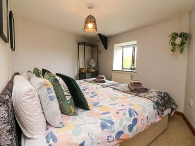 A bedroom with a bed covered in floral bedding and multiple pillows a window with plants and a hanging plant by the wall at Horseshoe Cottage in Hartland