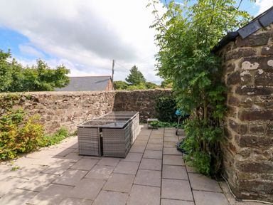 A paved outdoor patio area with wicker furniture a barbecue and stone walls at Horseshoe Cottage in Hartland