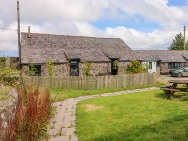 A stone cottage with slate roof and fenced garden beside a paved path with a wooden picnic table on grass at Horseshoe Cottage in Hartland