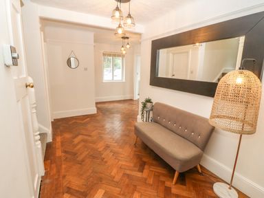 A hallway with a brown bench seat, a large rectangular mirror, a wicker floor lamp, a small side table with a plant, wooden parquet flooring, and a hanging round mirror at Afallon in Menai Bridge