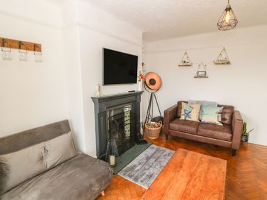 A living room with a brown leather sofa two gray chairs a wooden coffee table a black fireplace with a TV above a basket with firewood and wall shelves at Afallon in Menai Bridge