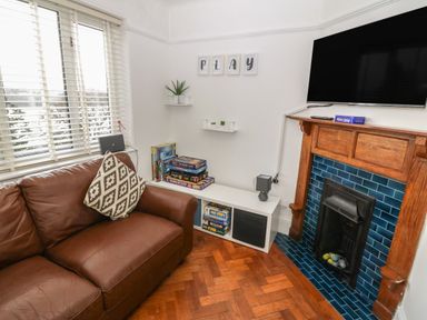 A corner of a living room with a brown leather sofa a white shelf holding board games and a speaker a wooden fireplace with blue tiles and a mounted TV at Afallon in Menai Bridge