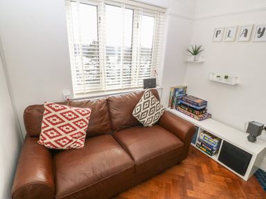 A brown leather sofa with two patterned cushions near a window with white blinds and a white shelving unit with board games and small plants at Afallon in Menai Bridge