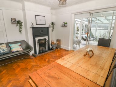 A dining room with a wooden table and black chairs a black fireplace and a black couch with cushions near sliding glass doors leading to a sunroom with chairs at Afallon Menai Bridge