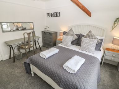 A bedroom with a bed covered in gray bedding and white towels on top a gray dresser and side tables with lamps and a desk with a chair and mirror at Afallon Menai Bridge
