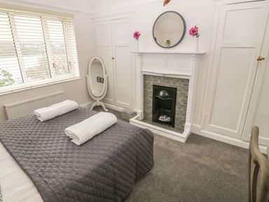 A bedroom with a bed covered with a gray quilt and towels a fireplace with a round mirror and flowers two white cupboards and a full length mirror at Afallon in Menai Bridge
