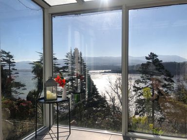A corner with glass walls overlooking trees and water with a small table holding a lantern and a vase with red flowers at Afallon in Menai Bridge