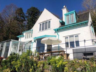 A white house with turquoise trims and a patio with a wooden table and umbrella at Afallon Menai Bridge