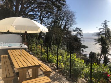 A wooden picnic table with benches and a white umbrella next to a hot tub overlooking a garden and a body of water at Afallon in Menai Bridge