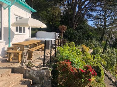 A garden patio with a wooden picnic table and umbrella next to a white house and hot tub surrounded by plants and trees at Afallon in Menai Bridge