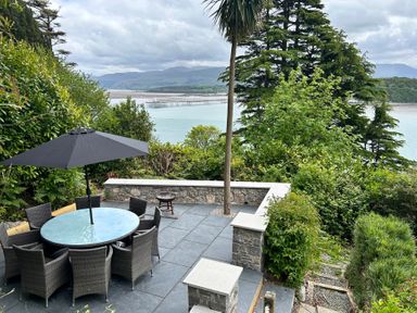 An outdoor patio with a round table and chairs under an umbrella overlooking a body of water with a pier and hills in the background at Afallon in Menai Bridge