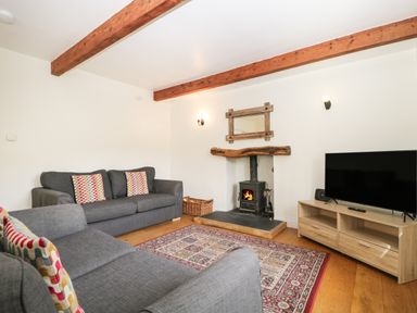 A living room with two gray sofas a wood burning stove a wooden TV stand with a flat screen TV a patterned rug and wooden ceiling beams at Glen Cottage in Troutbeck near Keswick