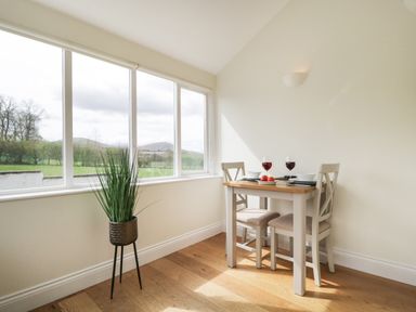 A dining area with a wooden floor a small table set for two and a tall plant by large windows at Glen Cottage in Troutbeck near Keswick