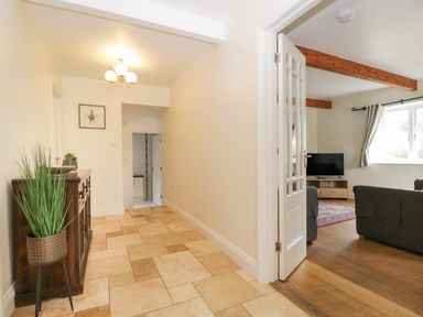 A hallway with a wooden cabinet a potted plant and a hanging light next to a living room with sofas a television and wooden beams at Glen Cottage in Troutbeck near Keswick