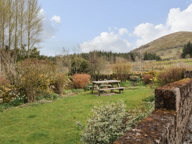 A garden with a wooden picnic table surrounded by bushes and trees with a hill and blue sky in the background at Glen Cottage in Troutbeck near Keswick