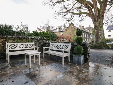 An outdoor seating area with two wooden benches and a small table on stone pavement near a stone wall and a large tree at Oak Cottage in Windermere