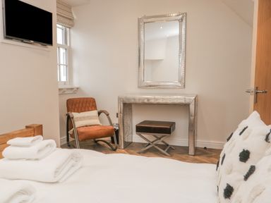 A bedroom with a bed with white linens and pillows a brown armchair a silver mirror and dressing table and a black stool at Oak Cottage in Windermere
