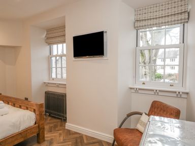A bedroom with wooden bed frame a wall-mounted television two windows with roman shades a radiator leather chair and metal table at Oak Cottage in Windermere