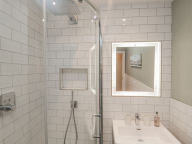A bathroom with a shower area with glass door white tiled walls a sink with two cups and a soap dispenser and an illuminated mirror at Oak Cottage in Windermere