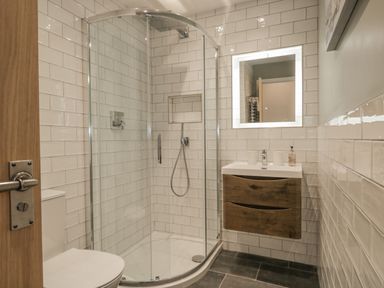 A bathroom with a curved glass shower enclosure a wall-mounted wooden vanity a mirror and a toilet at Oak Cottage in Windermere