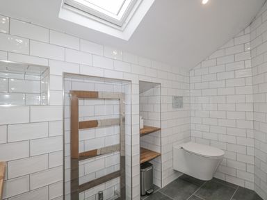 A bathroom with white tiled walls a wall mounted toilet a skylight and wooden shelves at Oak Cottage in Windermere
