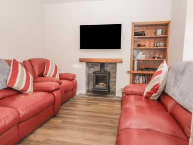 A living room with red leather sofas a wall-mounted TV a fireplace and a wooden shelving unit at Oak Cottage in Windermere