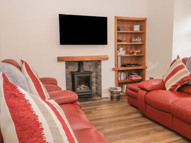 A living room with red leather sofas facing a wall-mounted TV and a fireplace with a wooden mantel and a wooden shelf unit at Oak Cottage in Windermere