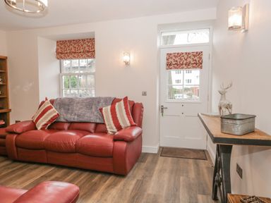 A living room with a red leather sofa two striped cushions a wooden side table with decorations and a white door with a window at Oak Cottage in Windermere