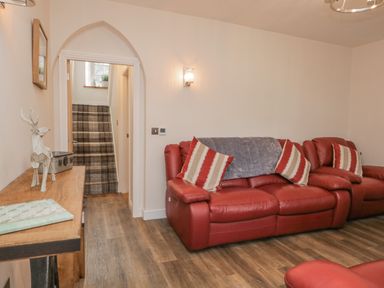 A room with a red leather sofa with striped cushions a wooden console table with a decorative deer a stairway with plaid carpet and wooden flooring at Oak Cottage in Windermere