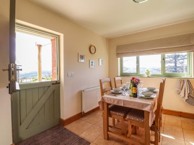 A dining area with a wooden table set for four near a half open green door and a window with cloth blinds at Barcud Cottage in Golden Grove near Llandeilo