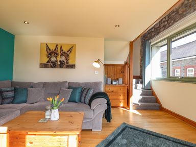 A living room with a gray sofa a wooden coffee table with flowers a wooden chest of drawers and stairs by a window at Barcud Cottage in Golden Grove near Llandeilo