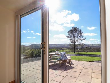 A patio with a wooden picnic table and a blanket on a chair overlooking a grassy area and hills at Barcud Cottage in Golden Grove near Llandeilo