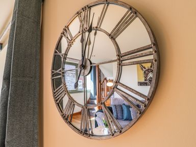 A wall clock with roman numerals reflecting a living area with a sofa cushions painting and staircase at Barcud Cottage in Golden Grove near Llandeilo