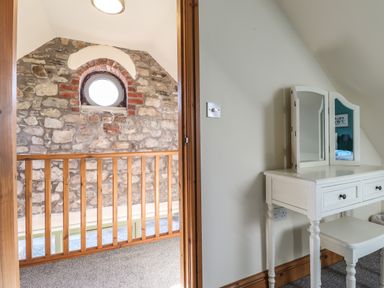 A dressing table with a tri-fold mirror and stool next to a wooden railing and a stone wall with a round window at Barcud Cottage Golden Grove near Llandeilo