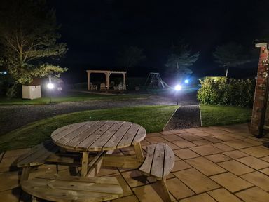 A nighttime outdoor area with a wooden picnic table on a tiled patio a gravel path leading to a pergola with chairs and a swing set at Barcud Cottage in Golden Grove near Llandeilo