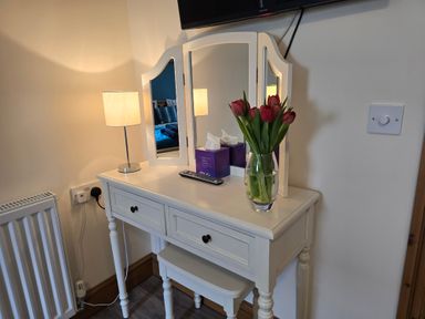 A dressing table with a mirror a lamp a remote a tissue box and a vase with flowers at Barcud Cottage in Golden Grove near Llandeilo