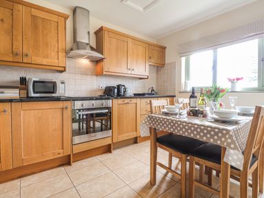A kitchen with wooden cabinets a stove microwave a dining table with tableware and flowers at Gwennol Cottage in Golden Grove near Llandeilo