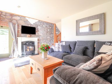 A living room with a gray sofa and pillows a wooden coffee table with flowers a fireplace with a mounted tv and a stone wall at Gwennol Cottage Golden Grove near Llandeilo