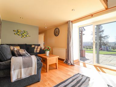 A living room with a dark sofa, wooden coffee table with flowers, wall clock, and sliding glass door at Gwennol Cottage in Golden Grove near Llandeilo