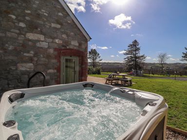 An outdoor hot tub next to a stone building with a picnic table and children's play area in the background at Gwennol Cottage Golden Grove near Llandeilo