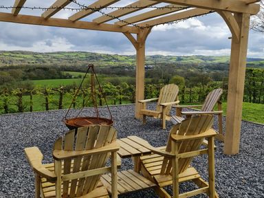 An outdoor seating area with wooden chairs and a fire pit under a wooden pergola overlooking green hills at Gwennol Cottage in Golden Grove near Llandeilo